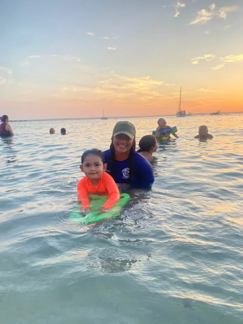 Nanda Paz, instructora de natación, dando clase a una niña con tabla en el mar al atardecer